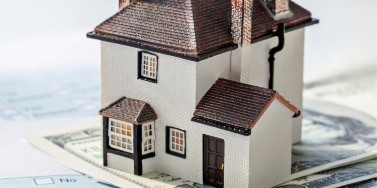 A small model house sits on top of dollar bills and mortgage documents, symbolizing home loans and financial decisions